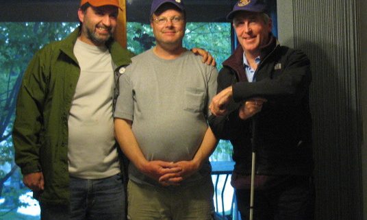 Three men standing indoors, wearing hats.