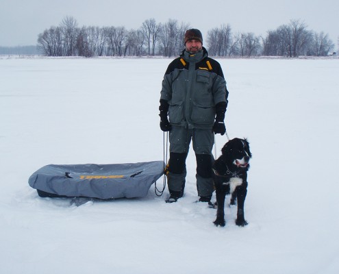 Man in winter gear with a dog on snowy landscape beside a sled.