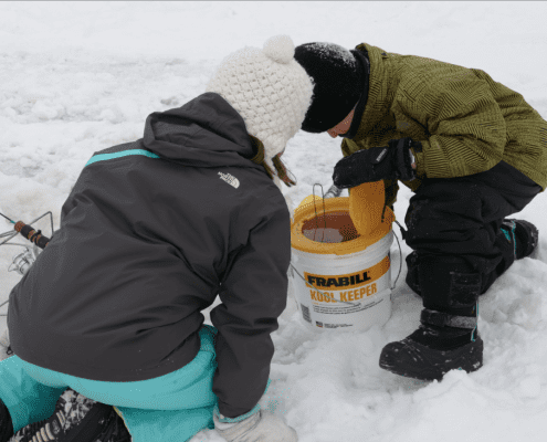 Two children playing in the snow near a colorful bucket.