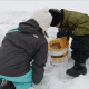 Two children playing in the snow near a colorful bucket.
