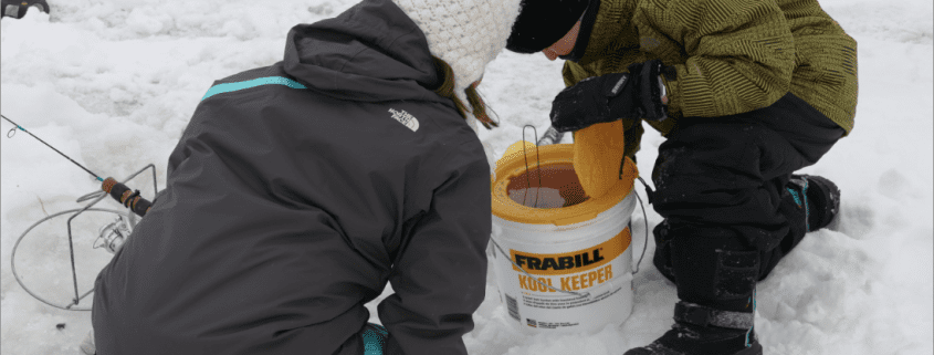 Two children playing in the snow near a colorful bucket.