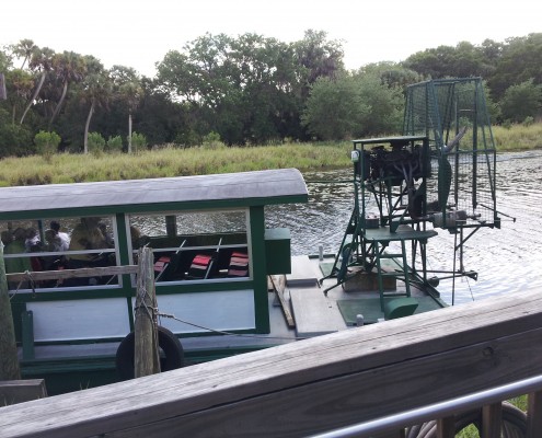 A sleek black speedboat docked by a wooden pier near a grassy shoreline.