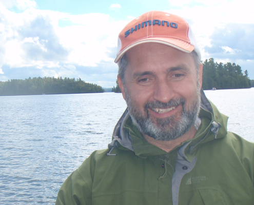 Man holding a fish by a lake with mountains in the background.
