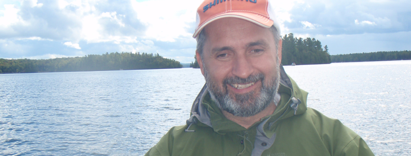 Man holding a fish by a lake with mountains in the background.