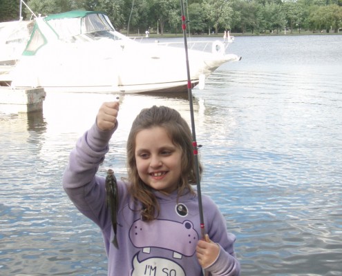 A smiling girl holding a fishing rod near a lake with boats.