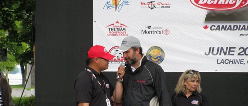 Two men speaking at an event, one wearing a red cap and the other a black cap.