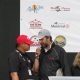 Two men speaking at an event, one wearing a red cap and the other a black cap.