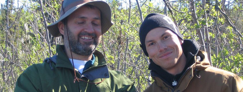 Two people outdoors, one holding a rifle, both looking intently in the same direction.