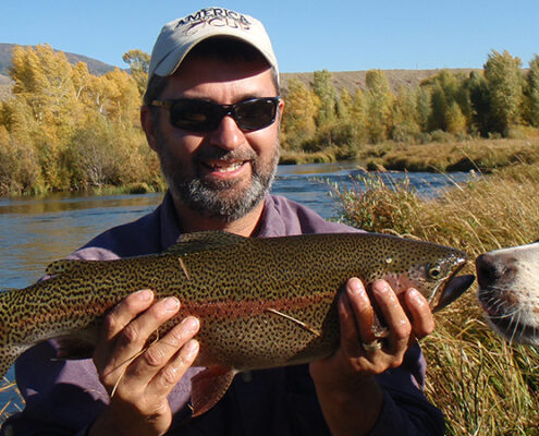 Man smiling and holding a large fish outdoors near a lake.