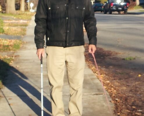 Man walking a small dog on a leash along a leaf-covered sidewalk.