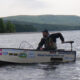 Person fishing on a small boat in a calm lake with hills in the background.