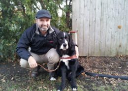 Man crouching with a black and white dog outside near a wooden fence.