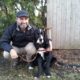 Man crouching with a black and white dog outside near a wooden fence.