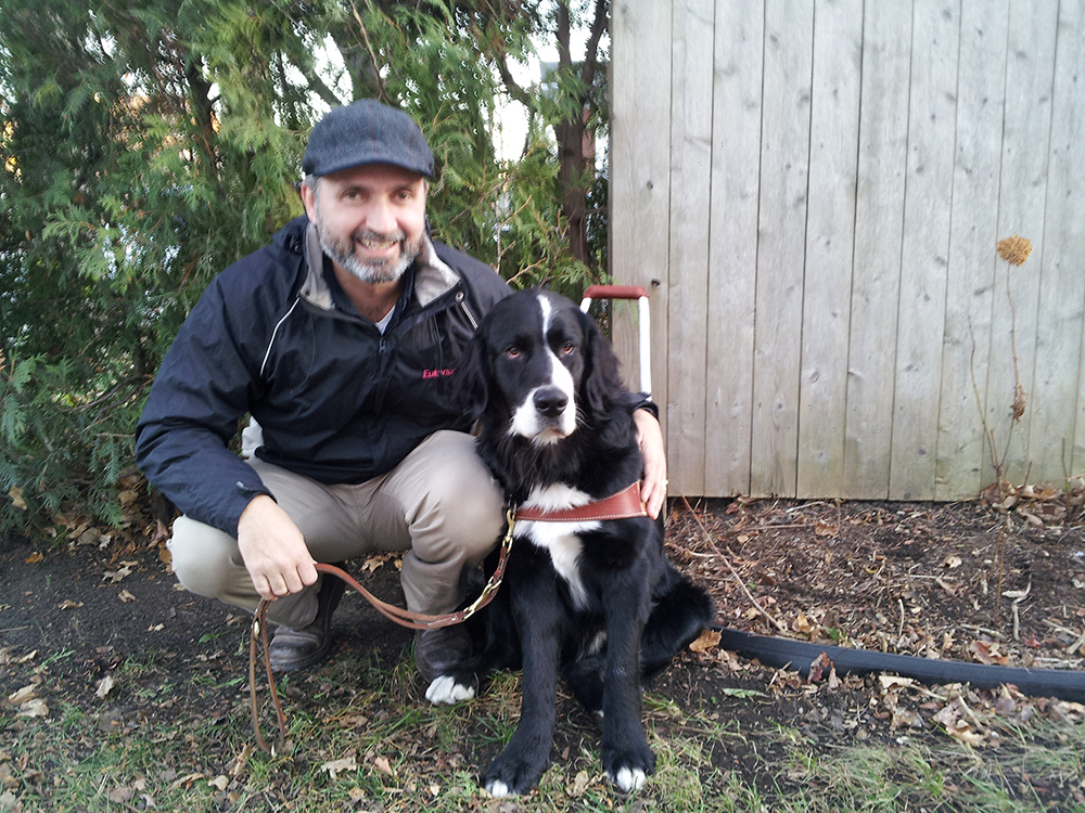 Man crouching with a black and white dog outside near a wooden fence.