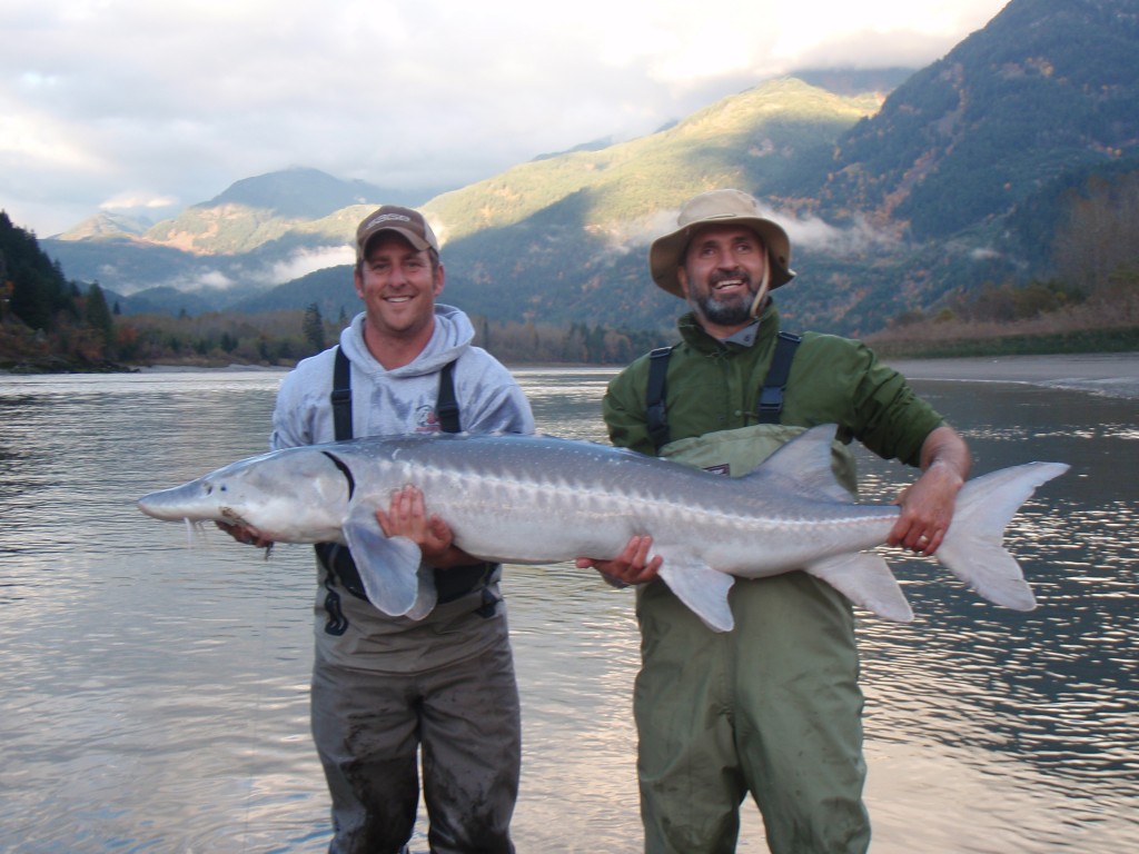 Two men proudly hold large fish by a river with mountains in the background.