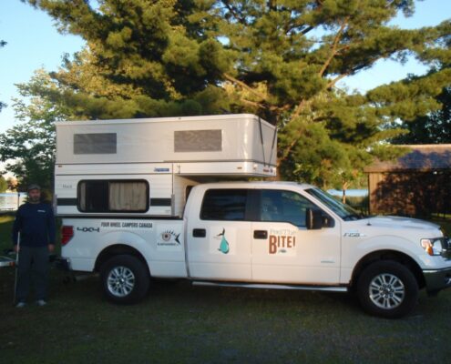 A white pickup truck towing a camper trailer in a wooded area during sunset.