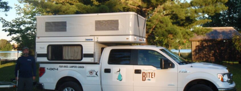 A white pickup truck towing a camper trailer in a wooded area during sunset.