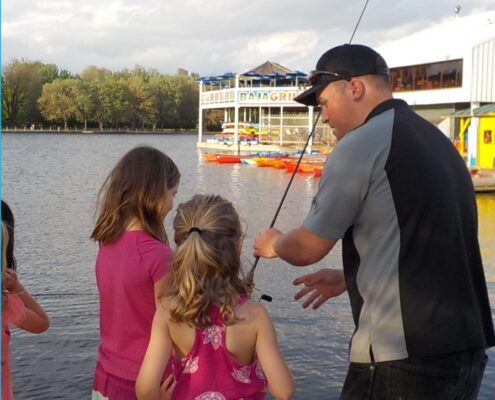 A man and two girls enjoying time near the water with boats in the background.