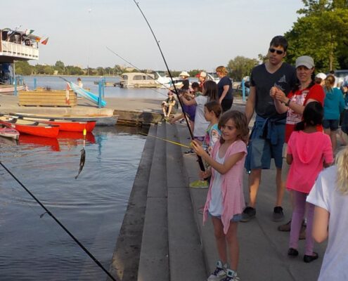 Children and adults feeding fish near a waterfront with boats.