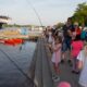 Children and adults feeding fish near a waterfront with boats.