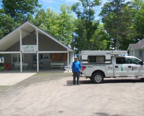 Man standing with his dog near a white truck and a fenced area in a wooded park.