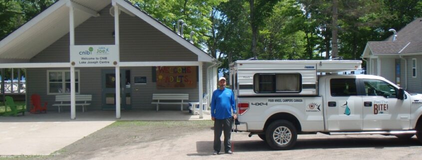 Man standing with his dog near a white truck and a fenced area in a wooded park.