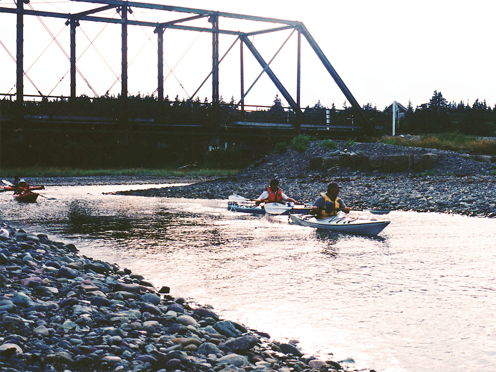 Two kayakers paddling on a calm river near a metal bridge.