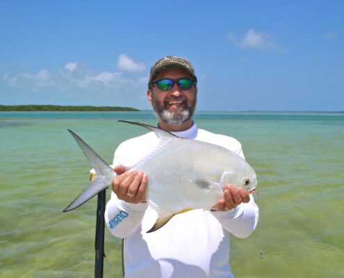 Man holding a fish on a sunny beach with clear blue water.